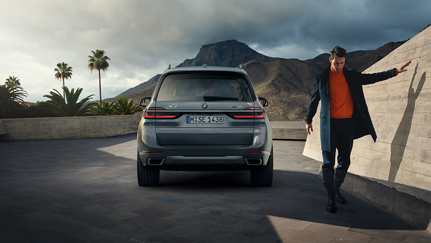 BMW X7, G07, LCI, perspetiva traseira com modelo à direita, pintura cinza Sparkling Copper metalizada